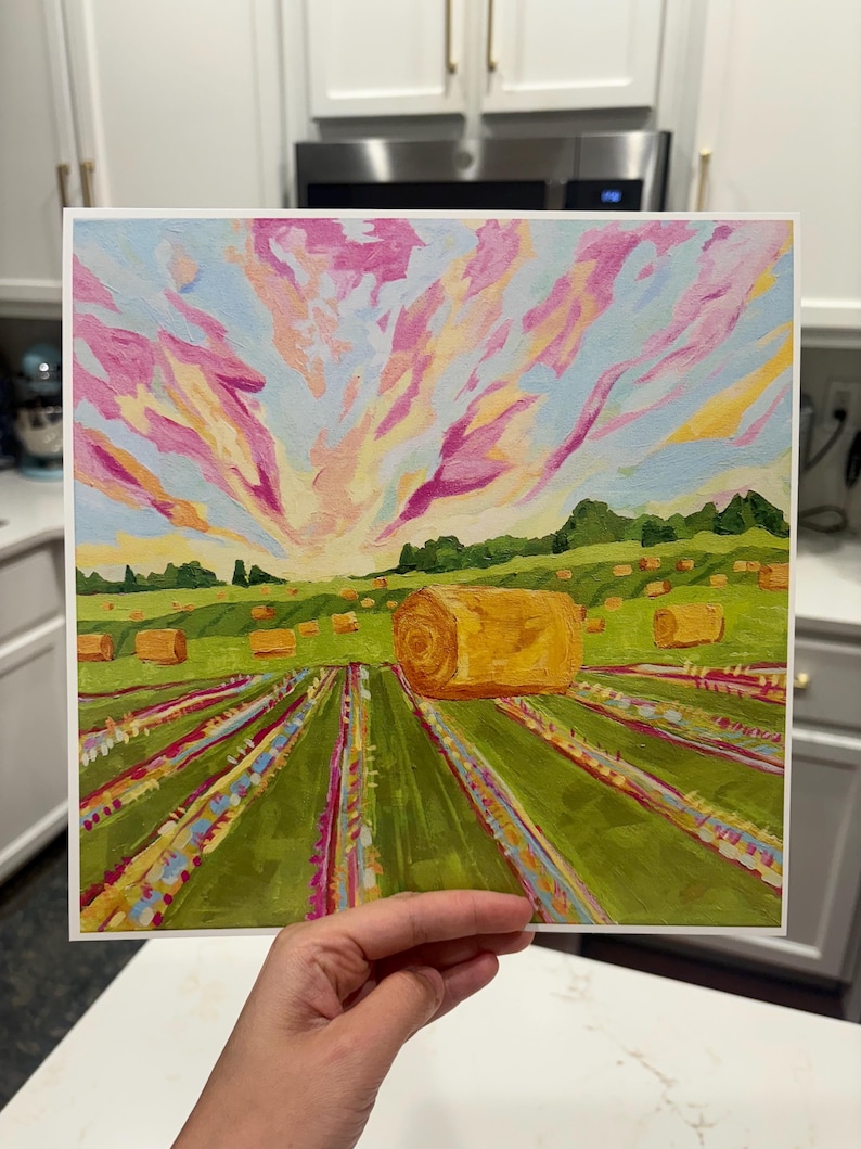 Puede incluir: Una impresi&oacute;n art&iacute;stica cuadrada que muestra un paisaje con un campo verde, varias pacas de heno doradas y un cielo vibrante lleno de nubes con tonos rosas, amarillos y azul claro. El primer plano muestra filas de flores coloridas.