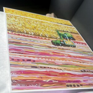 May include: A square art print featuring a painted landscape with a green combine harvester working in a golden yellow field. The foreground consists of horizontal bands of color in shades of pink, orange, red, and white, creating a textured effect.