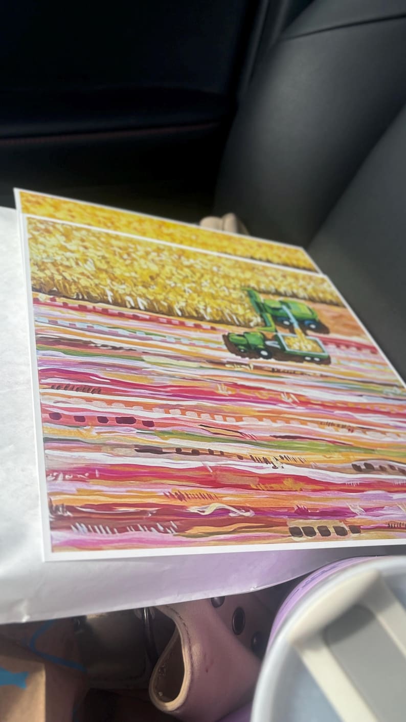 May include: A square art print featuring a painted landscape with a green combine harvester working in a golden yellow field. The foreground consists of horizontal bands of color in shades of pink, orange, red, and white, creating a textured effect.