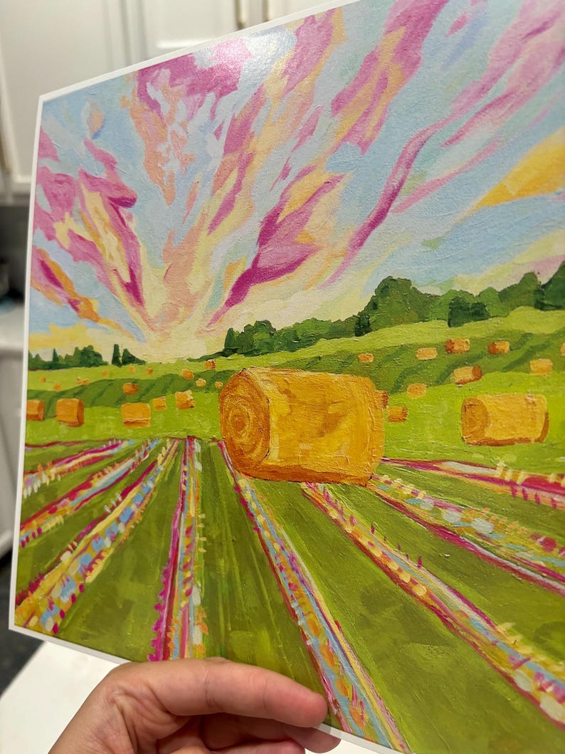 Puede incluir: Una impresi&oacute;n art&iacute;stica cuadrada que representa un paisaje rural con una gran paca de heno dorada en primer plano. El campo presenta hileras de hierba verde con acentos coloridos, que conducen a un horizonte con &aacute;rboles bajo un cielo rosa, amarillo y azul.