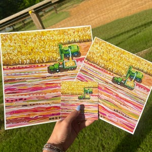 May include: Three square art prints featuring a painted scene of a green combine harvester working in a golden cornfield. The foreground displays horizontal bands of pink, red, orange, and white paint strokes, creating a textured, abstract landscape.