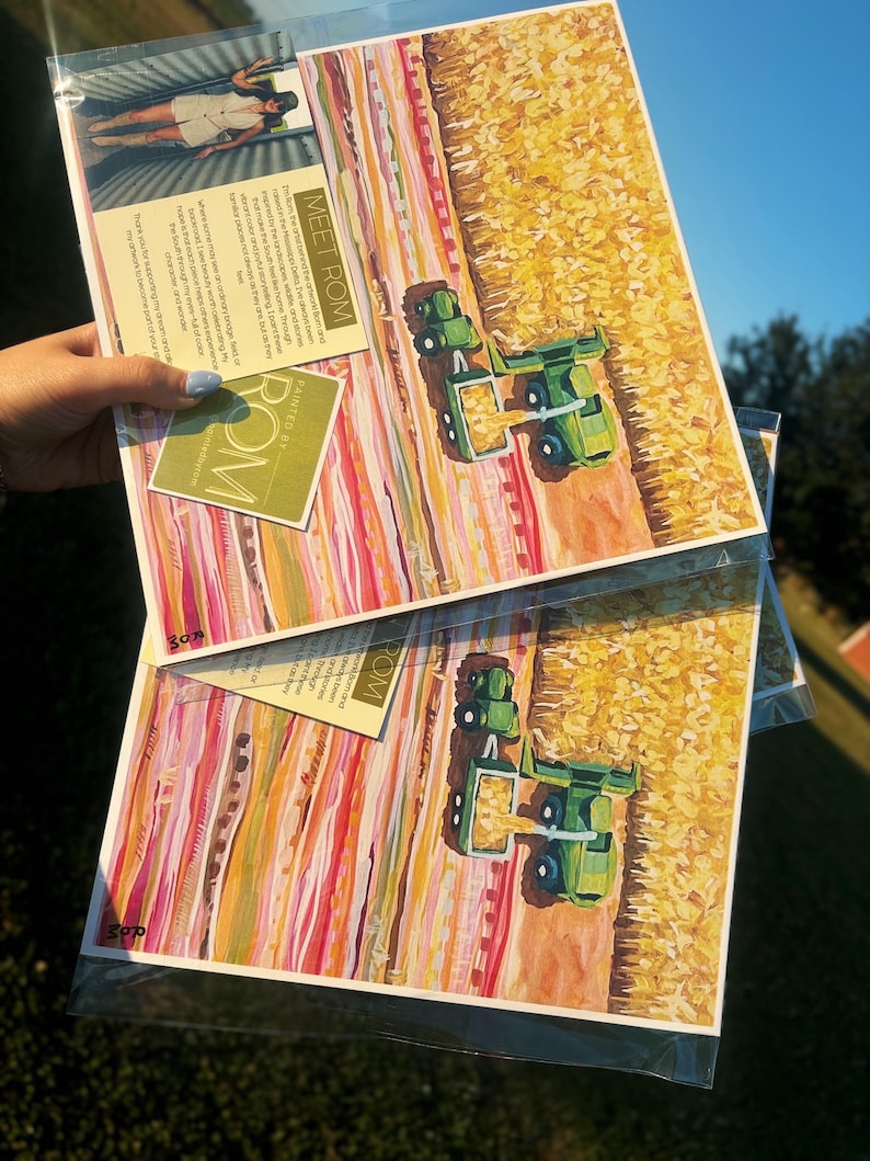 May include: Two identical art prints in clear plastic sleeves featuring a green tractor harvesting a yellow cornfield. The artwork includes horizontal bands of pink, orange, and red colors. A small card with the text "MEET ROM" is attached to the top.