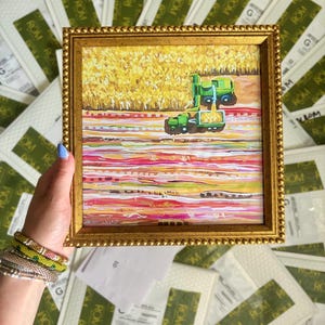 May include: A square painting in a gold beaded frame depicting a green combine harvester working in a yellow field. The lower portion of the artwork features horizontal bands of pink, white, and orange paint, creating a textured, abstract landscape.