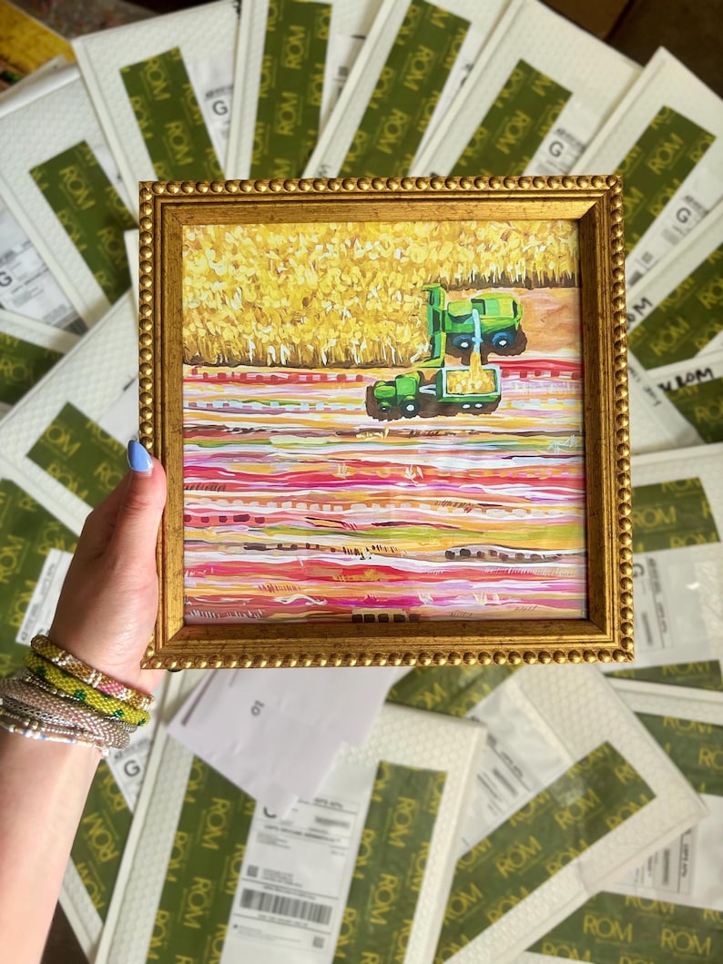 May include: A square painting in a gold beaded frame depicting a green combine harvester working in a yellow field. The lower portion of the artwork features horizontal bands of pink, white, and orange paint, creating a textured, abstract landscape.