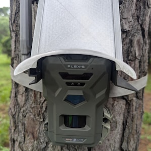 May include: A green and gray trail camera, secured to a tree with a strap. The camera has the text "FLEX-S" on the front and is protected by a white sun shield. The camera is designed for outdoor use.