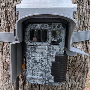 May include: A camouflaged trail camera with a gray protective housing is mounted on a tree. The camera has a black lens and a black and gray patterned front. A gray strap secures the camera to the tree.