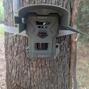 May include: A green and gray trail camera is mounted on a tree trunk. The camera has the text "FLEX-M" on the front and is secured with a strap. The camera is designed for outdoor use and wildlife monitoring.