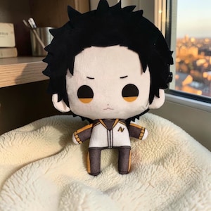 May include: A plush doll featuring a character with spiky black hair, large black eyes, and a pale face. The doll wears a white and dark grey jacket with orange accents and a small letter N embroidered on the chest, paired with matching dark grey trousers.