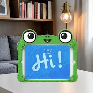 May include: A bright green tablet case shaped like a frog with large cartoon eyes and small frog feet at the corners. The screen displays the time 1:57 and the word Hi! in large white script against a blue background, framed by the green silicone.