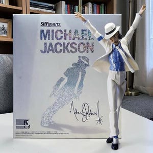 Può includere: Un action figure S.H.Figuarts di Michael Jackson in posa con le braccia alzate, che indossa un abito bianco, una camicia blu e un fedora. Accanto si trova la scatola originale con il nome Michael Jackson, una sagoma glitterata e una firma.