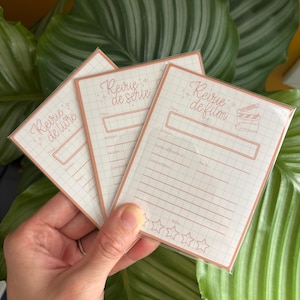 Peut inclure: Trois cartes rectangulaires en papier avec un motif quadrillé et des bordures couleur cuivre. Chaque carte présente des en-têtes en français dont 'Revue de livre', 'Revue de série' et 'Revue de film', avec des lignes pour notes et cinq étoiles vides.