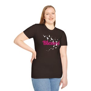 May include: A dark brown t-shirt featuring the pink word "Blessed" and a design of white birds in flight. The t-shirt is worn by a person wearing blue jeans.