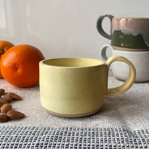 Può includere: Una tazza di ceramica gialla con un manico arrotondato è appoggiata su una superficie strutturata, accanto a due arance e una manciata di mandorle. Tre altre tazze sono impilate sullo sfondo, una verde, una bianca e una con bordo marrone e bianco. La scena è ambientata su un tessuto a motivi blu e bianco.