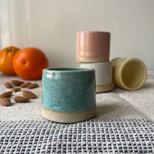 Puede incluir: Una colección de pequeñas tazas cilíndricas de cerámica en varios colores: turquesa, rosa, blanco y amarillo. Las tazas tienen una textura moteada y una base natural sin esmaltar. También se ven dos naranjas y almendras.