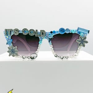 May include: Decorative sunglasses with a light blue frame, adorned with pearls, rhinestones, and snowflake accents. The text "IT'S COLD" is spelled out across the top of the frame. The lenses are a gradient purple.