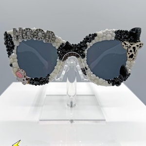 Puede incluir: Gafas de sol únicas con diseño de estampado de vaca. Las monturas están adornadas con cuentas blancas y negras, y la palabra "HOOD" está escrita con letras plateadas. Incluye dijes con temática de vaca y lentes oscuras.