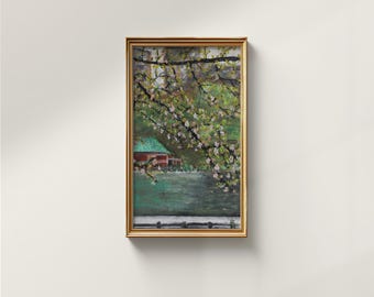 Pode incluir: Pintura de paisagem emoldurada com uma moldura dourada. A obra retrata um edifício vermelho com telhado verde perto de um corpo de água, emoldurado por galhos de árvores com flores rosa. A pintura está montada numa parede branca.
