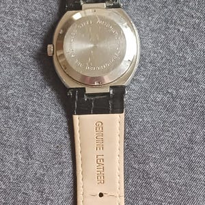 Puede incluir: Un reloj Kenneth Cole con una correa de cuero negro y una hebilla plateada. La parte trasera de la caja del reloj es plateada y tiene texto. La correa est&aacute; estampada con "Genuine Leather".