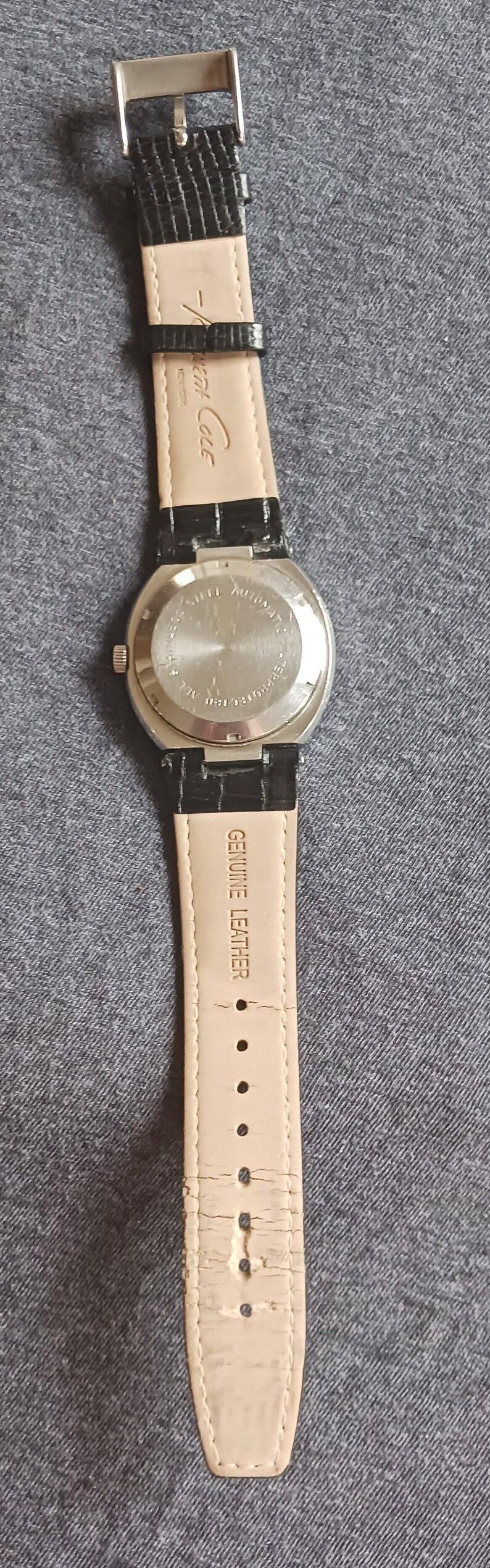 Puede incluir: Un reloj Kenneth Cole con una correa de cuero negro y una hebilla plateada. La parte trasera de la caja del reloj es plateada y tiene texto. La correa est&aacute; estampada con "Genuine Leather".