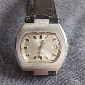 Puede incluir: Un reloj de pulsera plateado vintage con una esfera rectangular y una correa de cuero negro. La esfera es plateada con marcadores de hora y agujas negras. La esfera tiene la palabra "Stora" y "25 Rubis Automatic" impresas.