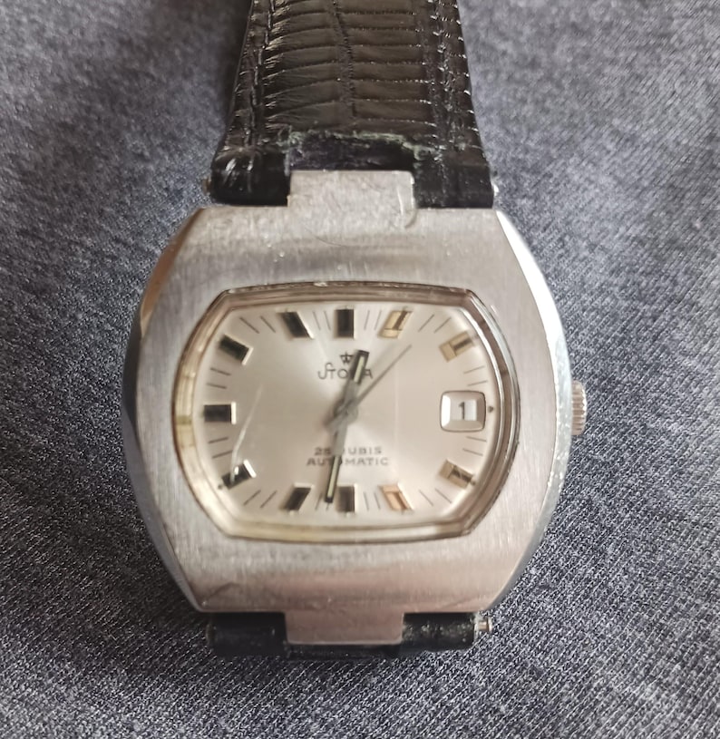 Puede incluir: Un reloj de pulsera plateado vintage con una esfera rectangular y una correa de cuero negro. La esfera es plateada con marcadores de hora y agujas negras. La esfera tiene la palabra "Stora" y "25 Rubis Automatic" impresas.