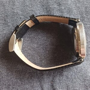 Puede incluir: Un reloj de pulsera plateado con una correa de cuero negro. La esfera del reloj es rectangular y la correa presenta un patr&oacute;n texturizado. El reloj est&aacute; sobre un fondo de tela gris.