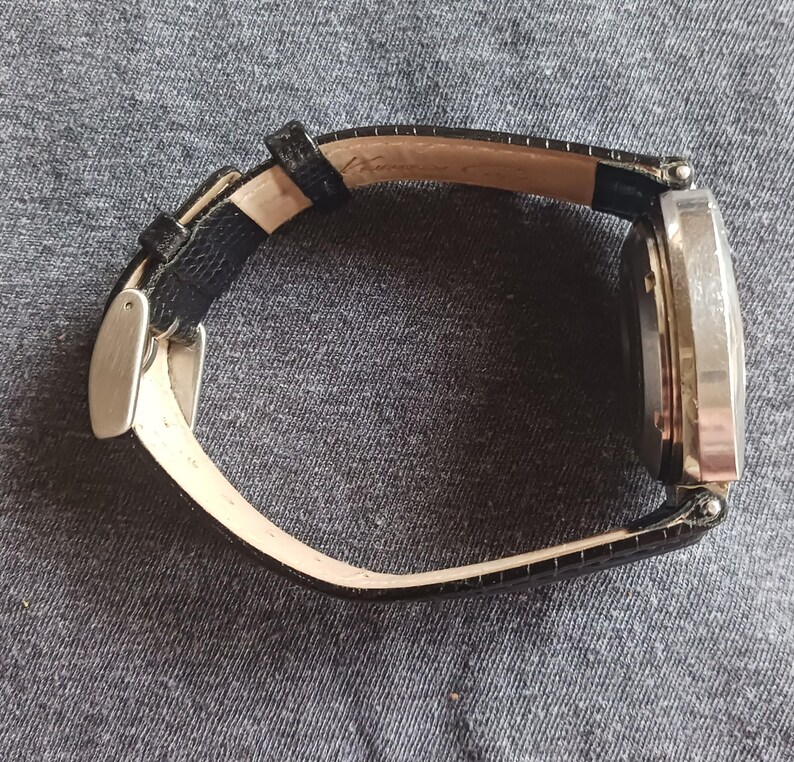 Puede incluir: Un reloj de pulsera plateado con una correa de cuero negro. La esfera del reloj es rectangular y la correa presenta un patr&oacute;n texturizado. El reloj est&aacute; sobre un fondo de tela gris.
