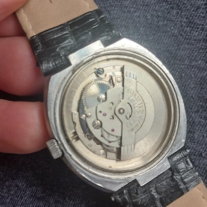 Puede incluir: Primer plano de la parte trasera de un reloj de pulsera vintage, que muestra el intrincado movimiento mec&aacute;nico. El reloj tiene una caja plateada, una correa de cuero negro y las palabras "17 JEWELS" son visibles. El reloj est&aacute; sobre una superficie de mezclilla.