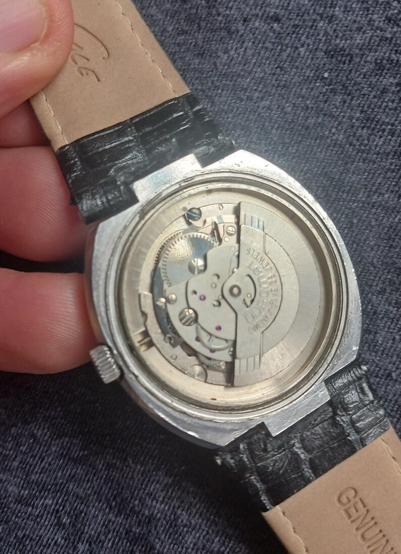 Puede incluir: Primer plano de la parte trasera de un reloj de pulsera vintage, que muestra el intrincado movimiento mec&aacute;nico. El reloj tiene una caja plateada, una correa de cuero negro y las palabras "17 JEWELS" son visibles. El reloj est&aacute; sobre una superficie de mezclilla.