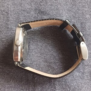 Puede incluir: Un reloj de pulsera plateado con una correa de cuero negro. El reloj tiene una esfera rectangular y un peque&ntilde;o bot&oacute;n redondo en el lateral. La correa se sujeta con una hebilla plateada.
