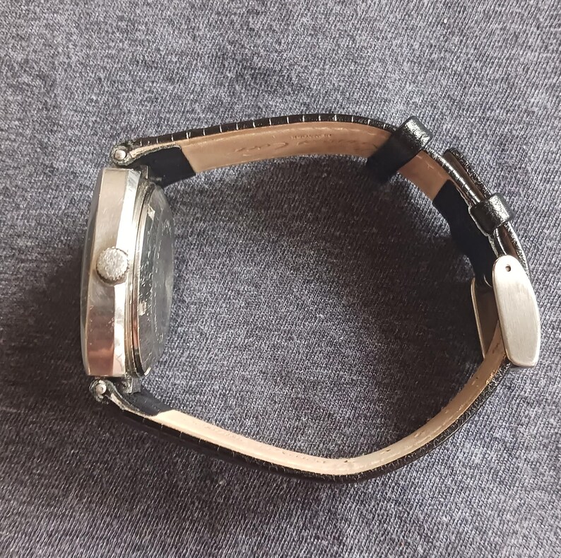 Puede incluir: Un reloj de pulsera plateado con una correa de cuero negro. El reloj tiene una esfera rectangular y un peque&ntilde;o bot&oacute;n redondo en el lateral. La correa se sujeta con una hebilla plateada.