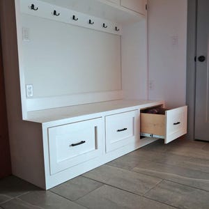 May include: White built-in storage unit with a bench, drawers, and hooks. The unit has a clean, modern design with black hardware. One drawer is open, revealing the interior. The unit is against a white wall and a gray tiled floor.