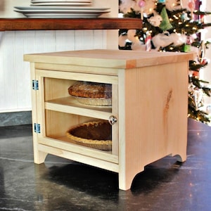 Może przedstawiać: Jasna drewniana szafka na ciasto ze szklanymi drzwiami, prezentująca dwa ciasta na półkach. Szafka ma prostokątny kształt z płaskim blatem i stoi na ciemnej powierzchni. Szafka ma około 61 cm wysokości.