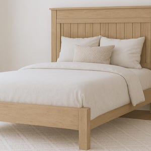 May include: A light-colored wooden bed frame with a matching headboard. The bed is made up with white bedding and pillows. A small decorative pillow is centered on the bed. A potted plant is to the right of the bed.