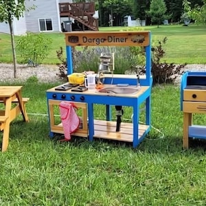 May include: A wooden play kitchen set with a blue frame and a wooden countertop. The set includes a sink, stove, and a sign that reads "Darga Diner". A small picnic table and a grill are also visible.