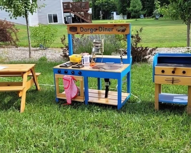 May include: A wooden play kitchen set with a blue frame and a wooden countertop. The set includes a sink, stove, and a sign that reads "Darga Diner". A small picnic table and a grill are also visible.