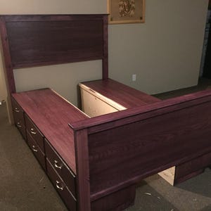 May include: A custom-built bed frame in a deep burgundy stain. The bed features a headboard and footboard, with built-in storage drawers on each side. The drawers have brass-colored hardware. The bed frame is made of wood and is set on a dark carpet.