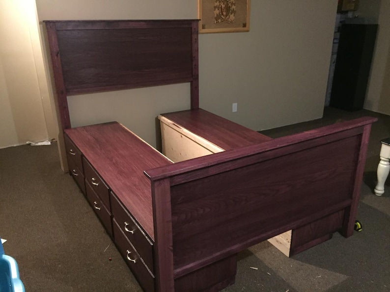 May include: A custom-built bed frame in a deep burgundy stain. The bed features a headboard and footboard, with built-in storage drawers on each side. The drawers have brass-colored hardware. The bed frame is made of wood and is set on a dark carpet.