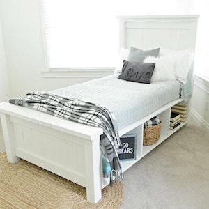Könnte beinhalten: Ein weißes Holzbett mit Kopfteil, hellgrauer Steppdecke und Kissen. Eine grau-weiß karierte Decke ist über dem Fußende des Bettes drapiert. Das Bett verfügt über einen integrierten Stauraum mit einem Korb und Büchern. Ein Schild liest "GO BEARS".