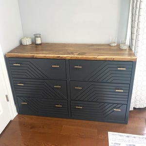 May include: A six-drawer dresser with a dark gray finish and geometric design on the drawer fronts. The top is made of stained wood. The dresser has gold-colored hardware and is placed against a gray wall.