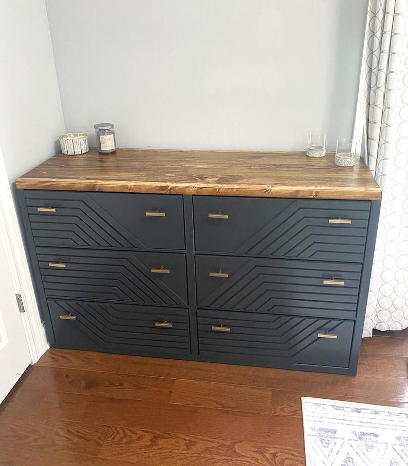 May include: A six-drawer dresser with a dark gray finish and geometric design on the drawer fronts. The top is made of stained wood. The dresser has gold-colored hardware and is placed against a gray wall.