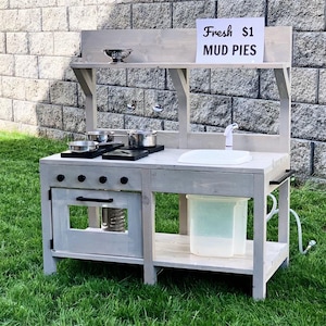 May include: A gray wooden play kitchen set with a sink, stove, and oven. The set includes pots, pans, and a sign that reads "Fresh $1 MUD PIES". The play kitchen is set up on a grassy lawn.