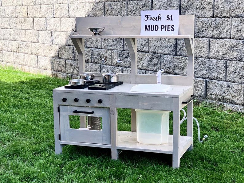 May include: A gray wooden play kitchen set with a sink, stove, and oven. The set includes pots, pans, and a sign that reads "Fresh $1 MUD PIES". The play kitchen is set up on a grassy lawn.