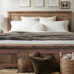 Può includere: Una struttura letto in legno rustico con testiera e pediera, dotata di materasso bianco, cuscini bianchi, un cuscino rotondo bianco e una coperta in pelliccia sintetica grigio scuro. Un cesto intrecciato con coperte arrotolate si trova ai piedi.