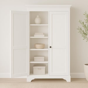 May include: A white wooden cabinet with two doors, one open, revealing shelves with decorative items. A vase, books, and boxes are visible. A potted plant with green leaves stands to the right. The cabinet is a storage solution.