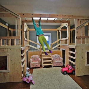 May include: A multi-level wooden playhouse with a climbing bar, and a child hanging from it. The playhouse has a light wood finish and includes a small pink toy car and two floral armchairs. The structure is indoors.