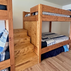 May include: A wooden bunk bed with a staircase. The bed frames are made of light brown wood. The bedding includes blue and white patterned quilts and solid blue sheets. The stairs are made of the same wood as the bed frame.