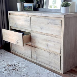 Może przedstawiać: Jasna drewniana komoda z sześcioma szufladami, z których jedna jest otwarta. Komoda ma naturalne wykończenie słojów drewna i jest umieszczona przy ścianie z szarą zasłoną. Przed komodą znajduje się dywan z kwiatowym wzorem.