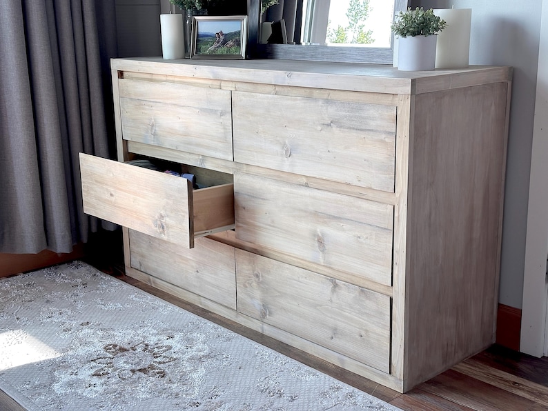 May include: A light-colored wooden dresser with six drawers, one of which is open. The dresser has a natural wood grain finish and is placed against a wall with a gray curtain. A rug with a floral pattern is in front of the dresser.