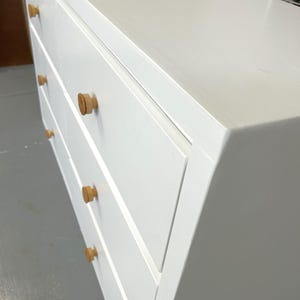 May include: A white dresser with four drawers and light wood knobs. The dresser is painted white and has a simple, modern design. The knobs are round and made of a light-colored wood.
